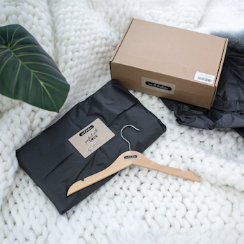 Schwarzes Paket mit einem Kleiderbügel und einer Notiz auf einem weißen, gestrickten Untergrund, daneben ein brauner Karton." 

(Note: The text is already in German and does not require translation. It is clear and fluent as it stands.)