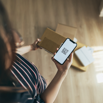 Eine Person steht neben mehreren Kartons und hält ein Smartphone mit einem QR-Code in der Hand.