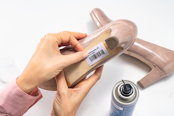 Eine Hand hält einen Schuh mit einem Preisschild, während daneben eine Spraydose steht. Der Schuh ist ein beiger Absatzschuh.