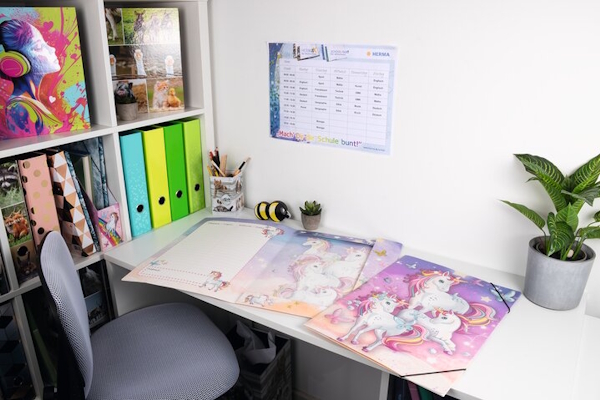 Ein Schreibtisch, geschmückt mit mehreren Einhörnern auf Postern, einem Wandkalender, bunten Ordnern und einer Topfpflanze.