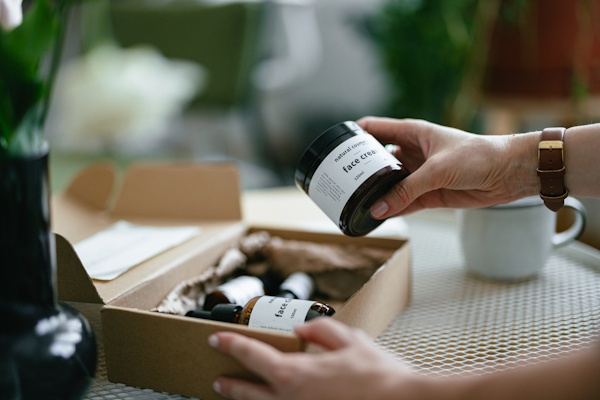 Eine Hand hält ein Glas mit einer Creme, während auf einem Tisch eine geöffnete Verpackung mit weiteren Produkten liegt.