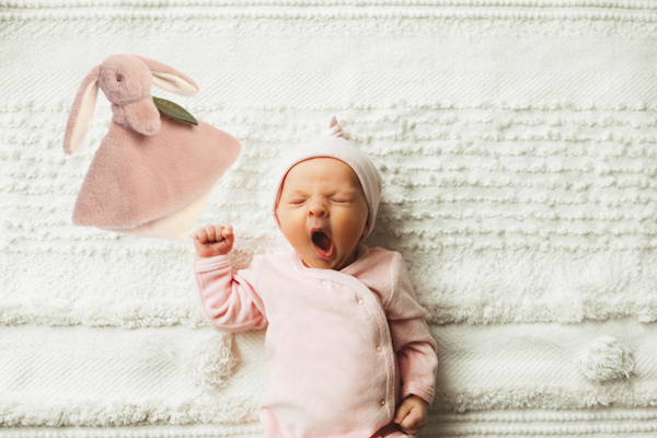 Ein schlafendes Baby in rosa Kleidung gähnt, während ein rosa Stoffhase neben ihm liegt. Das Baby trägt eine Mütze und liegt auf einer gestrickten Decke.