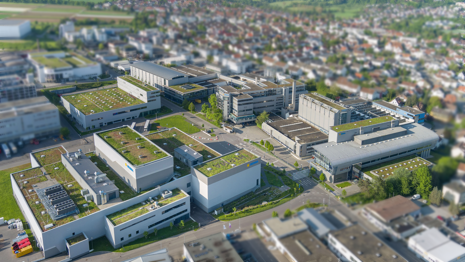 Luftaufnahme eines modernen Gewerbegebiets mit mehreren Gebäuden und begrünten Dächern, umgeben von einer städtischen Landschaft.