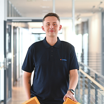 Ein junger Mann in einem dunkelblauen Poloshirt steht in einem modernen Büroflur und hält einen orangefarbenen Gegenstand in der Hand." 

(Note: The text is already in German and does not require translation. It is clear and fluent as it stands.)