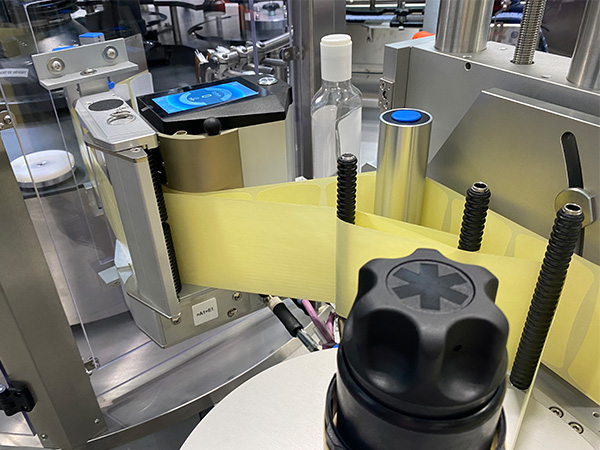 Ein Etikettiergerät mit gelbem Etikett, das auf Flaschen aufgebracht wird, befindet sich in einer Produktionsumgebung.