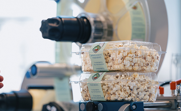 Zwei transparente Behälter mit Popcorn sind übereinander gestapelt, während im Hintergrund eine Popcornmaschine zu sehen ist.
