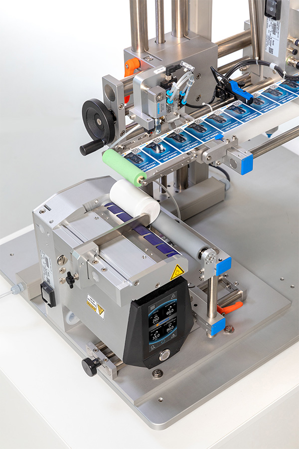Automatische Etikettiermaschine mit mehreren Rollen, die Etiketten auf einer Förderbandanordnung anbringt. Steuerungseinheit mit Display sichtbar.