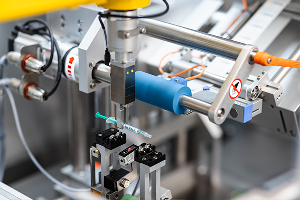 Automatisierte Maschine mit einem Greifarm, der eine Spritze hält, umgeben von verschiedenen mechanischen Komponenten und Sensoren.