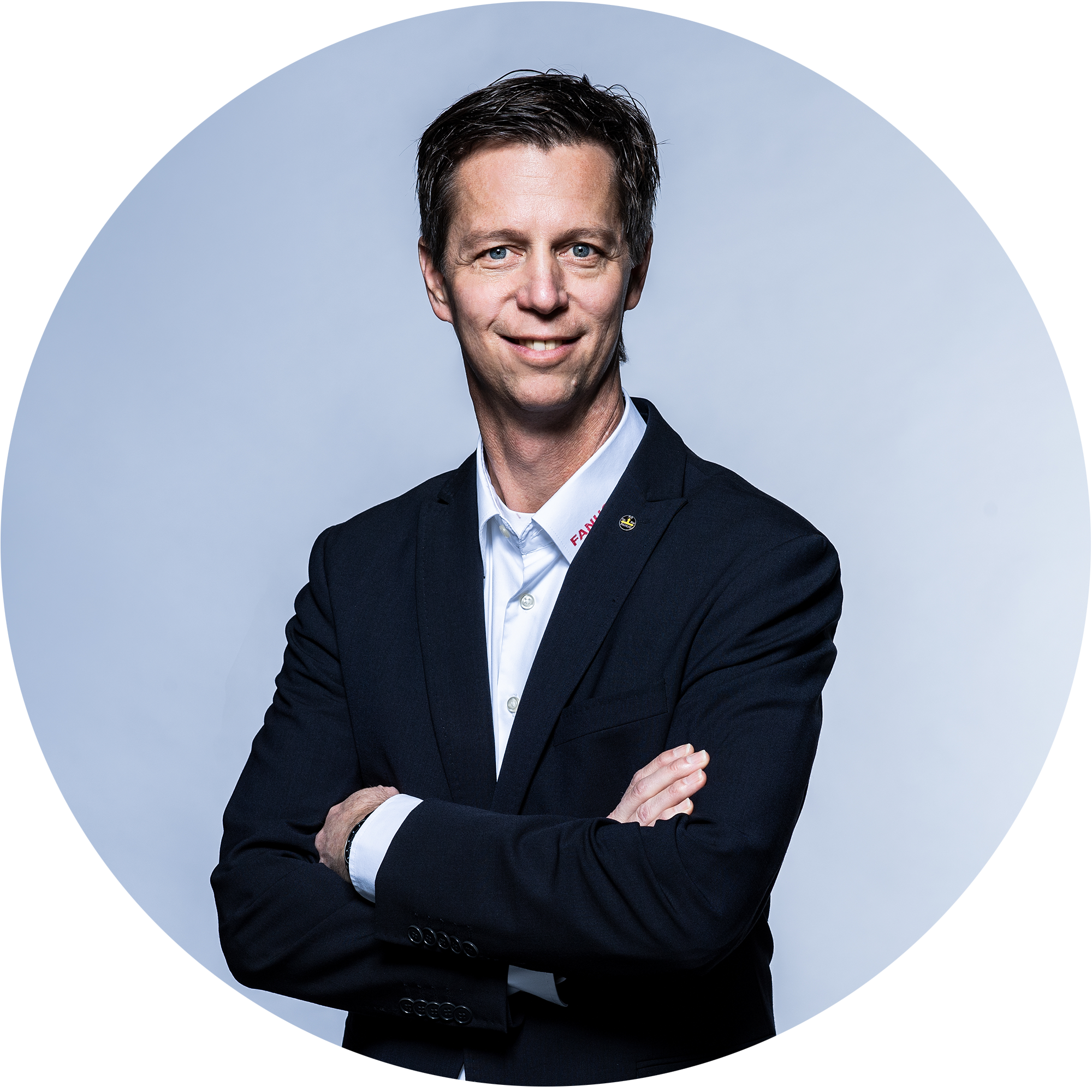 Ein Mann in einem schwarzen Anzug mit verschränkten Armen lächelt in die Kamera. Der Hintergrund ist hell und neutral." 

(Note: The text is already in German and does not require translation. It is clear and fluent as it is.)