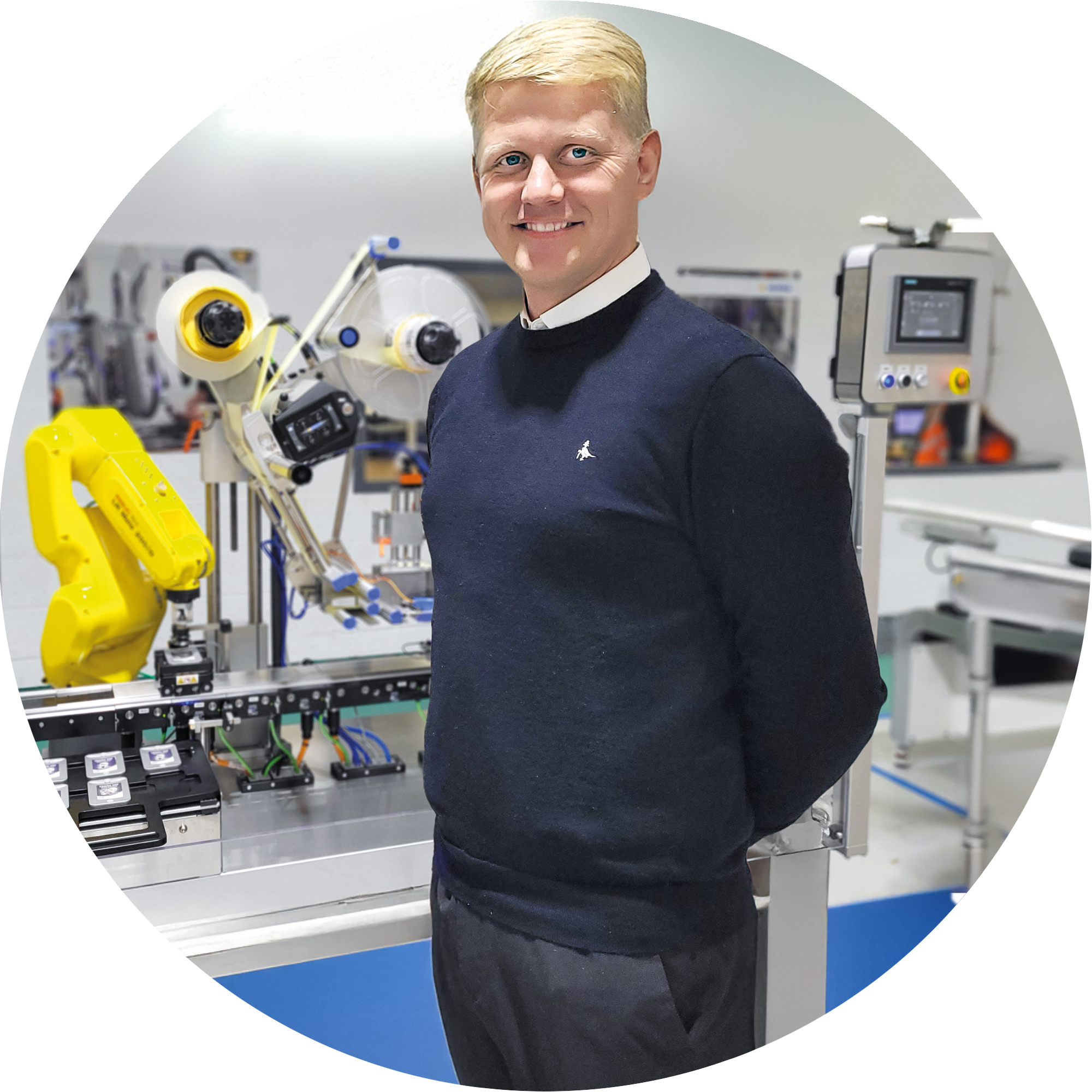 Ein Mann in einem schwarzen Pullover steht neben einer automatisierten Produktionslinie, die mit Robotern und technischen Geräten ausgestattet ist.