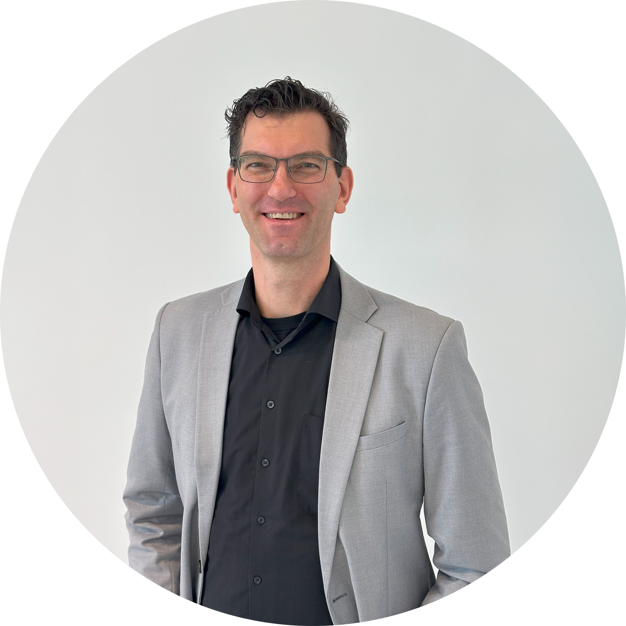 Ein Mann mit Brille trägt einen grauen Blazer über einem schwarzen Hemd und lächelt vor einem hellen Hintergrund." 

(Note: The text is already in German and does not require translation. It is clear and fluent as it is.)