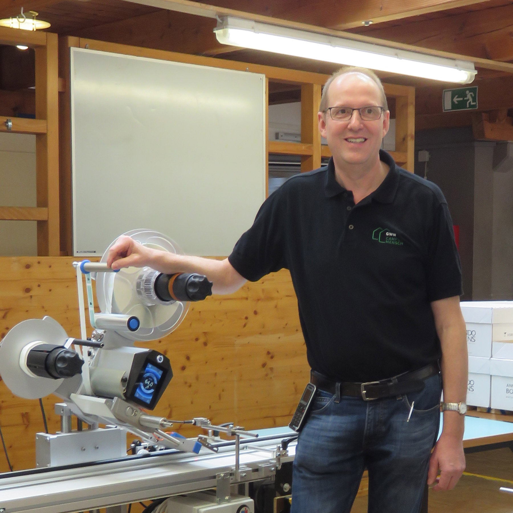 Ein Mann steht neben einer Maschine in einer Werkstatt, lächelt und trägt ein schwarzes T-Shirt mit einem Logo.