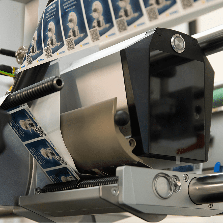 Eine Etikettiermaschine mit einer Rolle Etiketten, die QR-Codes und Bilder zeigt, steht bereit zur Verwendung in einer Produktionsumgebung.