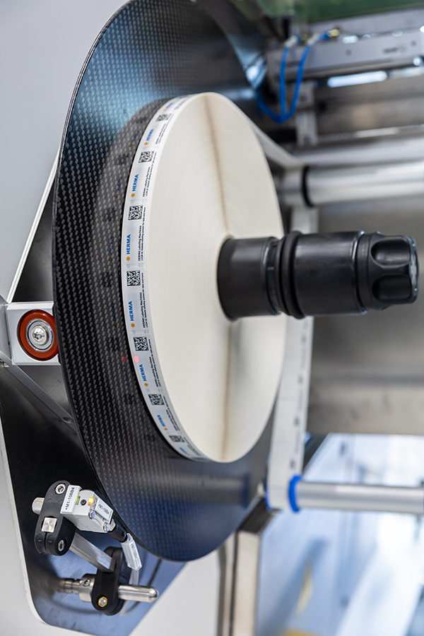 Eine große, runde Carbonrolle mit einem zentralen schwarzen Halter, umgeben von technischen Komponenten und Sensoren.
