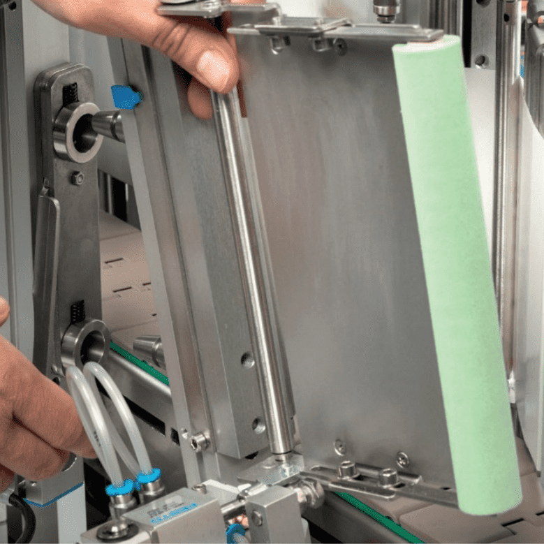 Eine Hand justiert eine mechanische Vorrichtung mit Metallteilen und einem grünen Element. Die Umgebung zeigt industrielle Ausrüstung.