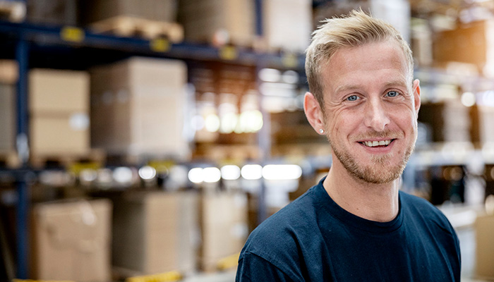Ein lächelnder Mann mit blonden Haaren steht in einem Lager mit Regalen voller Kartons im Hintergrund.