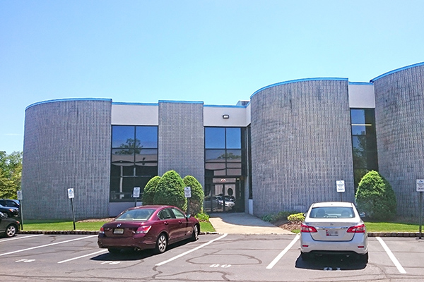 Moderne Gebäude mit grauer Fassade, großen Fenstern und zwei parkenden Autos im Vordergrund. Grüne Sträucher flankieren den Eingang.