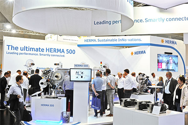 Messestand von HERMA mit der Präsentation des HERMA 500. Besucher interagieren mit den ausgestellten Maschinen und Informationen.