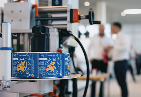 Im Vordergrund ist eine Verpackungsmaschine zu sehen, die bunte Etiketten auf Dosen anbringt, während im Hintergrund Personen miteinander sprechen.