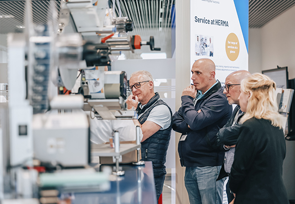 Vier Personen verfolgen aufmerksam eine Maschinenpräsentation auf einer Messe, während Informationen über den Service von HERMA eingeblendet werden.