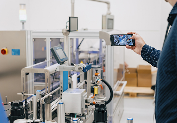 Eine Person hält ein Smartphone und filmt eine automatisierte Produktionsanlage in einer Fabrikumgebung.