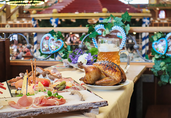 Ein Tisch, der mit einer Auswahl an Wurst, Käse und Gemüse gedeckt ist, dazu ein knuspriges Hähnchen und ein großes Bierglas in einer festlichen Atmosphäre.