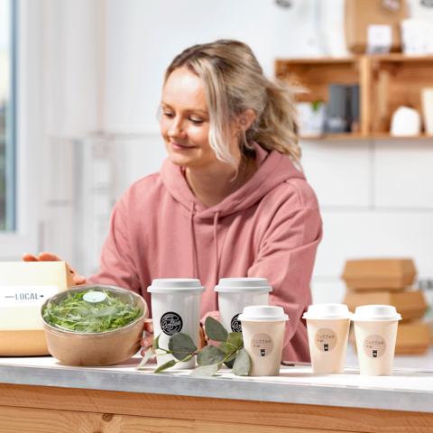 Eine Frau in einem rosa Hoodie bereitet gesunde Gerichte vor, darunter eine Schüssel Salat und mehrere Kaffeetassen auf einem Tisch.