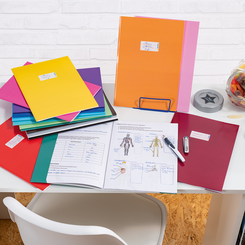 Bunte Ordner und Unterlagen auf einem Schreibtisch, daneben ein Glas mit Süßigkeiten und Bürobedarf wie einen Hefter und einen Stift.