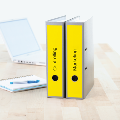 Zwei Ordner mit den Beschriftungen "Controlling" und "Marketing" stehen auf einem Tisch neben einem Laptop und einem Notizblock.