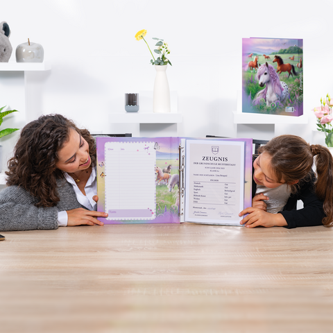 Zwei Mädchen sitzen am Tisch und schauen sich ein offenes Schulzeugnis in einem bunten Ordner mit Einhorn-Motiv an.