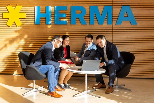 Blick auf grüne Baumkronen und eine Besprechung von drei Personen in einem modernen Büro mit dem Schriftzug "HERMA" im Hintergrund.