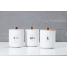Drei weiße Behälter mit Holzdeckeln, beschriftet mit "Zucker", "Kaffee" und "Tee", auf einem hellen Tisch vor einer grauen Wand.
