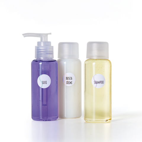 Drei Flaschen mit Pflegeprodukten: eine lila Flasche mit Pumpe, eine weiße Flasche und eine gelbe Flasche, beschriftet mit "Shampoo" und "Dusche Creme".
