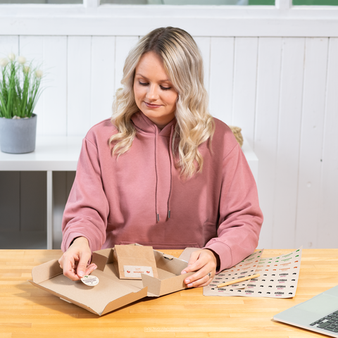 Eine Person mit lockigen, blonden Haaren sitzt an einem Tisch und arbeitet an einer Verpackung, während sie sich Aufkleber ansieht.