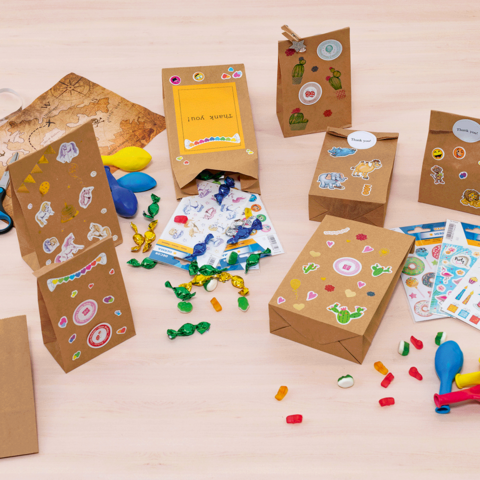 Bunte Geschenktüten und Bastelmaterialien auf einem Tisch, darunter Aufkleber, Luftballons, Scheren und Süßigkeiten." 

(Note: The text is already in German and does not require translation. It is clear and fluent as it stands.)