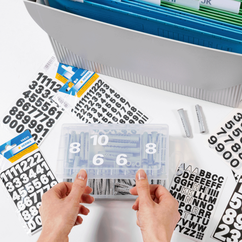 Hände halten eine transparente Box mit Zahlen und Buchstabenaufklebern, umgeben von weiteren Aufklebern und einem Ordner im Hintergrund.