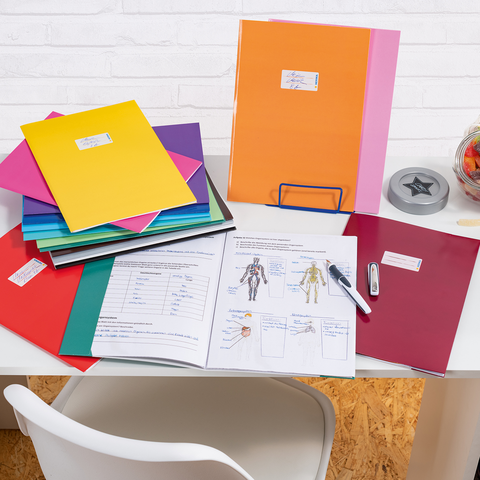 Bunte Ordner und Dokumente auf einem Schreibtisch, daneben ein Stift, ein Tacker und ein Glas mit Süßigkeiten.