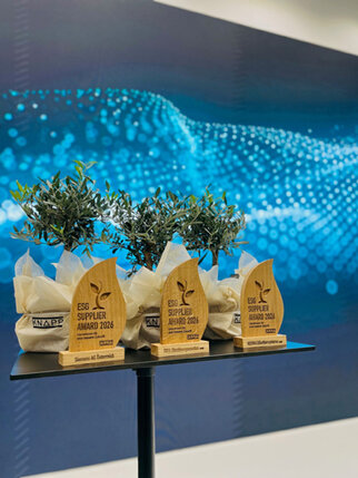 Trophäen des ESG Supplier Award 2026 stehen auf einem Tisch, umgeben von kleinen Pflanzen in Töpfen, vor einem blauen, digitalen Hintergrund.
