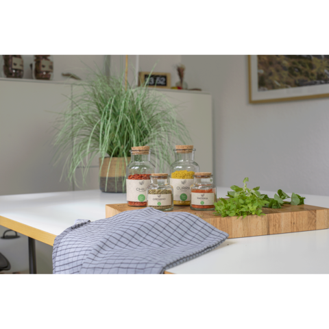 Gläser mit Gewürzen auf einem Tisch, daneben frische Kräuter und ein Küchentuch. Im Hintergrund eine Pflanze in einem Topf.