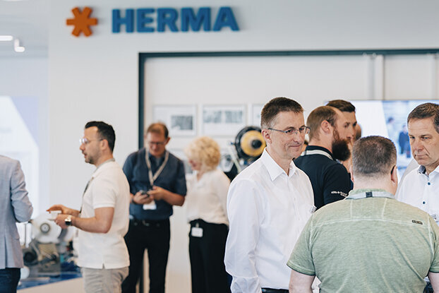 Menschen in formeller Kleidung stehen in einem modernen Raum, diskutieren und interagieren, während im Hintergrund ein HERMA-Schild zu sehen ist.