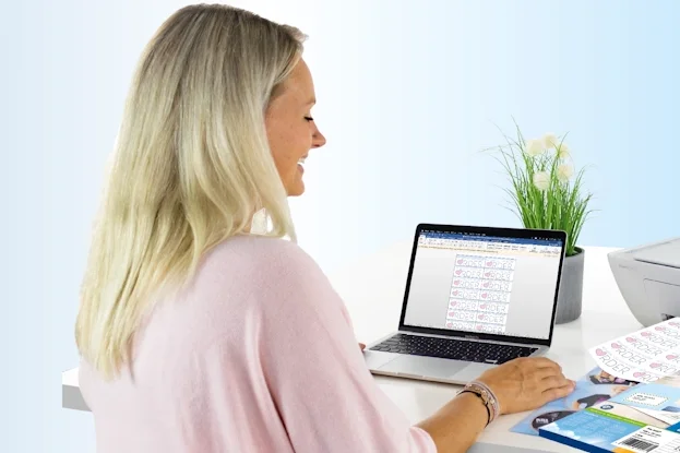 Eine Frau sitzt an einem Tisch mit einem Laptop und druckt Etiketten. Neben ihr steht ein Drucker sowie einige leere Etikettenblätter.