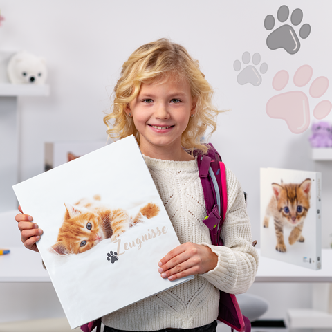 Ein Mädchen mit blonden Locken hält einen Ordner mit einem Katzenbild. Im Hintergrund sind weitere Ordner und Spielzeug zu sehen."
(Note: The text is already in German and does not require translation. It is clear and fluent as it stands.)