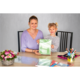 Eine Frau und ein Mädchen sitzen an einem Tisch und halten ein grünes Heft. Auf dem Tisch liegen verschiedene Schreibmaterialien und Blumen.