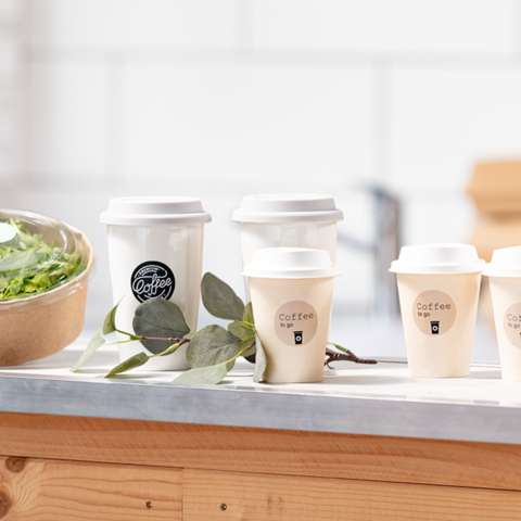 Auf einem Tisch stehen mehrere Kaffeebecher, neben einer Schüssel mit Salat und frischen grünen Blättern.