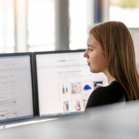 Eine Frau sitzt vor zwei Monitoren und betrachtet Inhalte auf einem Bildschirm, während sie konzentriert arbeitet.