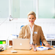 Eine Frau mit blonden Haaren sitzt an einem Schreibtisch, lächelt und trägt einen beigen Blazer. Vor ihr steht ein Laptop und Schreibutensilien.
