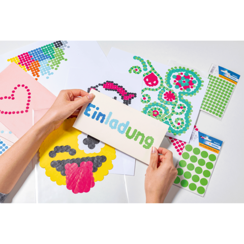 Hände halten eine Einladungskarte mit dem Wort "Einladung" in bunten Buchstaben. Um sie herum liegen Bastelmaterialien und Aufkleber.