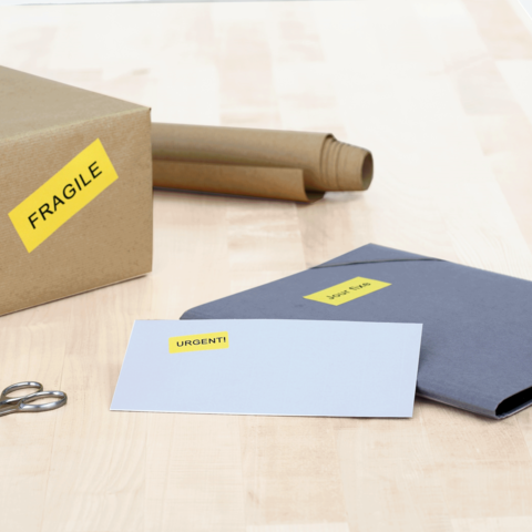 Ein Karton mit dem Aufkleber "FRAGILE", daneben ein graues Kuvert mit dem Aufkleber "URGENT!" und eine Schere auf einem Tisch.