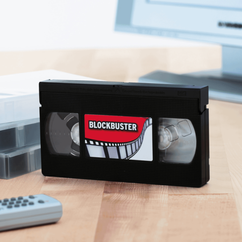 Eine schwarze VHS-Kassette mit einem Blockbuster-Etikett liegt auf einem Tisch neben einer Fernbedienung und einer transparenten Kassette.