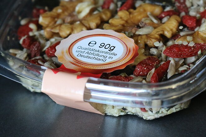 Eine transparente Schale mit einer Mischung aus Nüssen, Samen und getrockneten Früchten, etikettiert mit Nährwertinformationen.